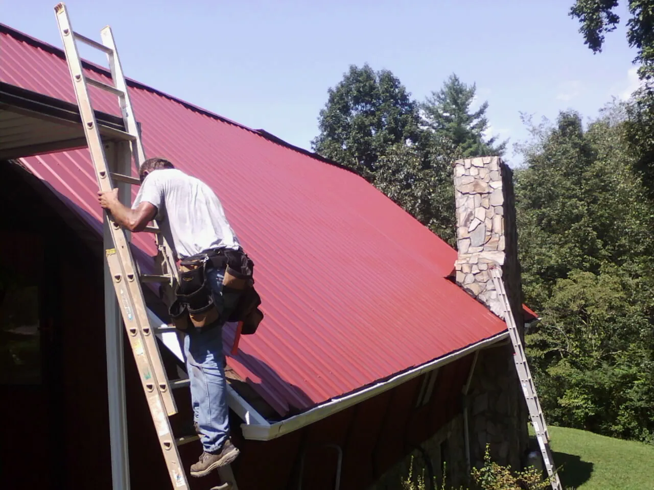 Roofer on a ladder beside a red metal roof and stone chimney — Soffit & Fascia Repair by Roofing Brothers in Tremont